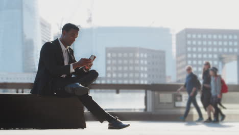 Millennial-black-businessman-wearing-a-suit-and-a-white-shirt-sitting-on-the-River-Thames-embankment-using-his-smartphone,-full-length