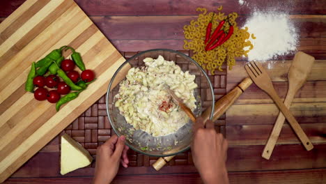 una vista dall'alto della marinatura dei maccheroni con ingredienti, maccheroni marinati nella ciotola, maccheroni crudi, cucchiaio di legno, spatola di legno, farina, mattarello, un pezzo di formaggio congelato e peperoncini sul tavolo