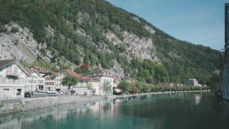 Pedestrians-riding-their-bikes-throughout-calming-village-on-mountainside-alongside-relaxing-river-early-morning-in-Interlaken,-Switzerland-in-Europe,-wide-view