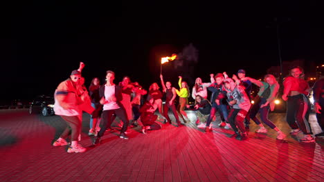 group of teenagers standing in half-circle dance hip-hop. young man in the middle holding a lit torch. night square backdrop.