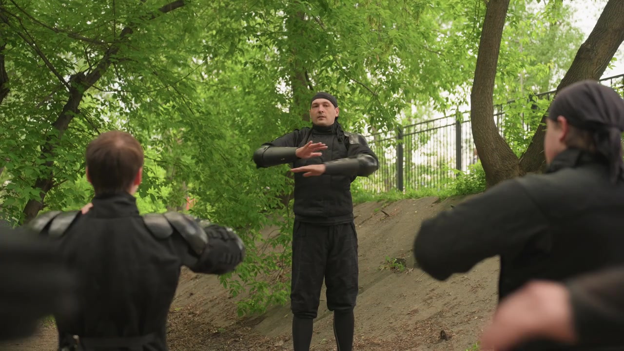 Primo piano di un uomo che dimostra tecniche di arti marziali ad altri due uomini in equipaggiamento protettivo nero all'aperto. Il dimostratore usa movimenti delle mani mentre i partecipanti imitano le sue azioni, tutti indossando