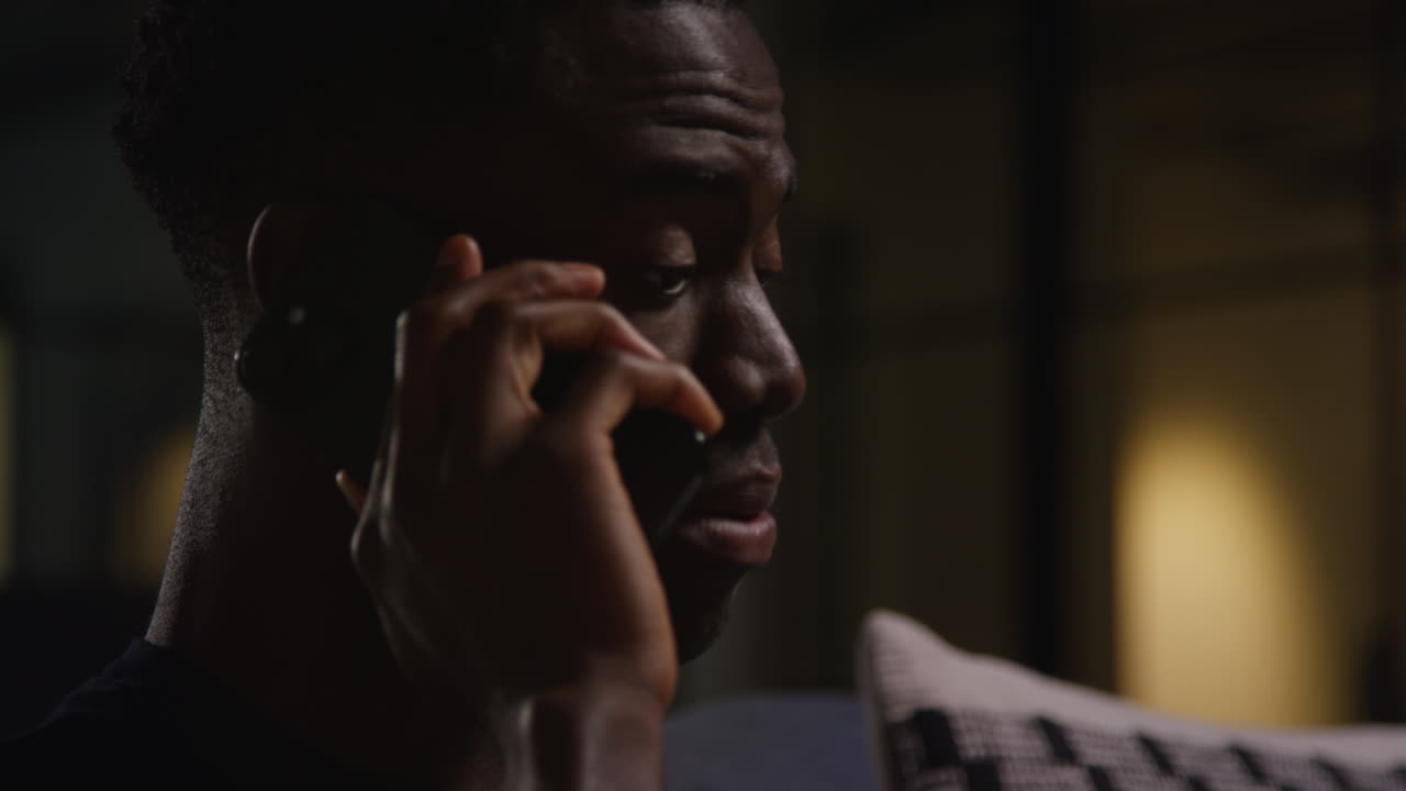 Close Up Of Unhappy And Depressed Young Man Sitting On Sofa At Home Making Call On Mobile Phone At Night 1