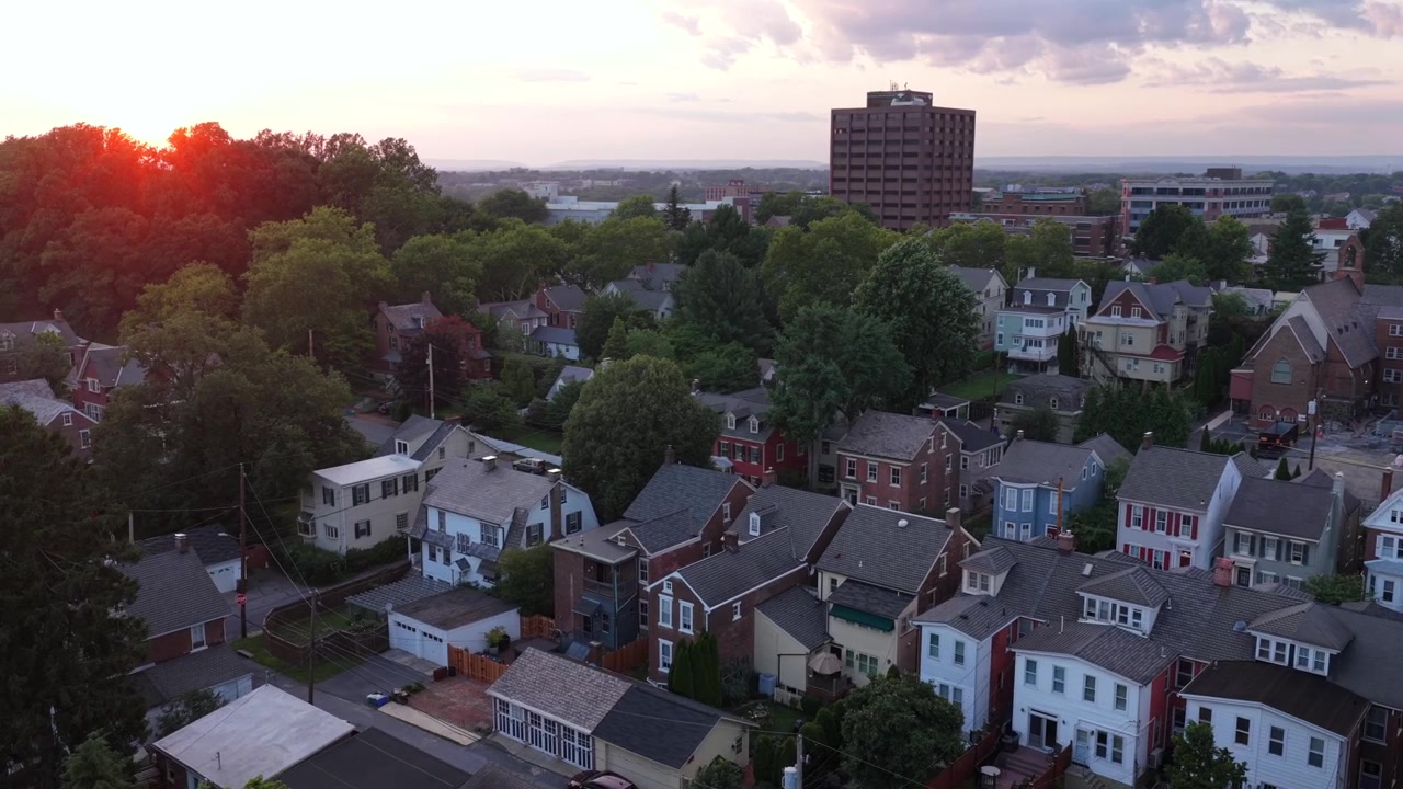 Victorian homes and brick rowhouses line dense tree-covered streets, tall office tower rises above quiet residential rooftops at golden sunset in Bethlehem, Pennsylvania. Aerial drone wide shot