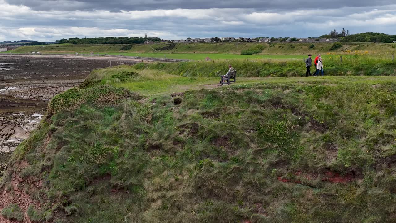 Drone glides over eroded red sandstone cliff, grassy headland, and seaside path beneath cloudy sky