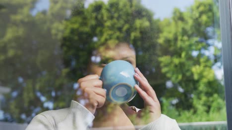 video of relaxed plus size african american woman drinking coffee and looking outside window