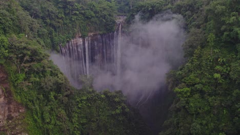 tumpak sewu waterfall hidden behind mist with semeru volcano, aerial