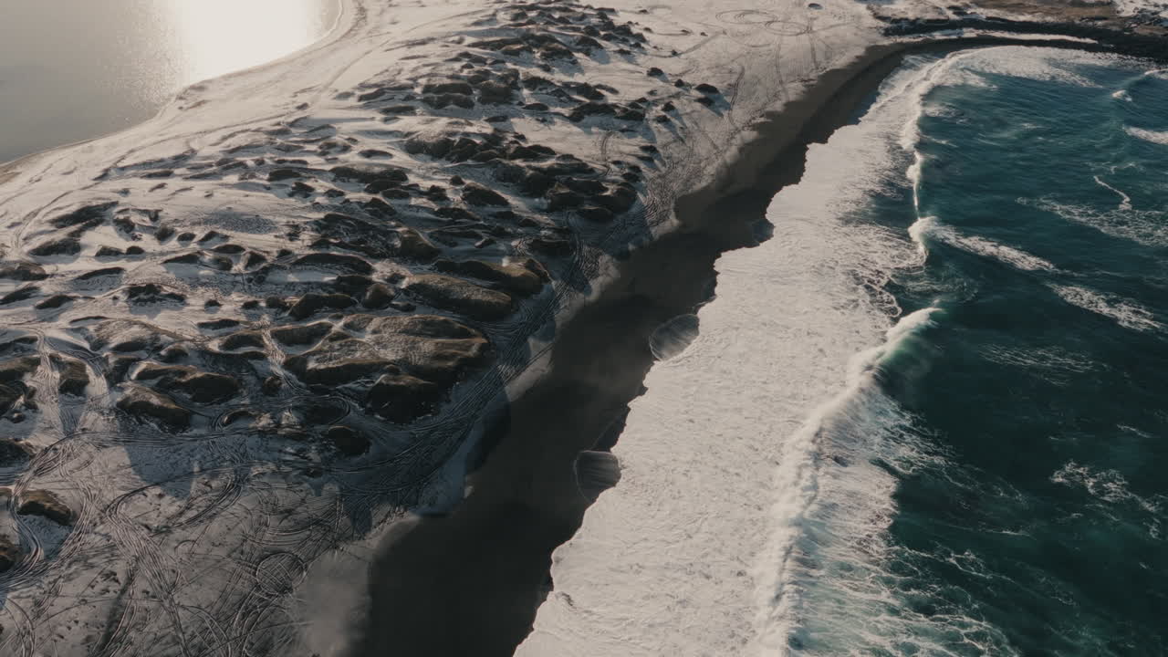 Drone top-down aerial view of ocean waves breaking onto a black sand beach alongside a rocky volcanic coastline in Iceland during winter. Coastal nature and Nordic wilderness concept