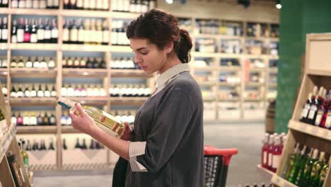 young beautiful brunette girl picks a bottle of wine from the shelf to read a label on it