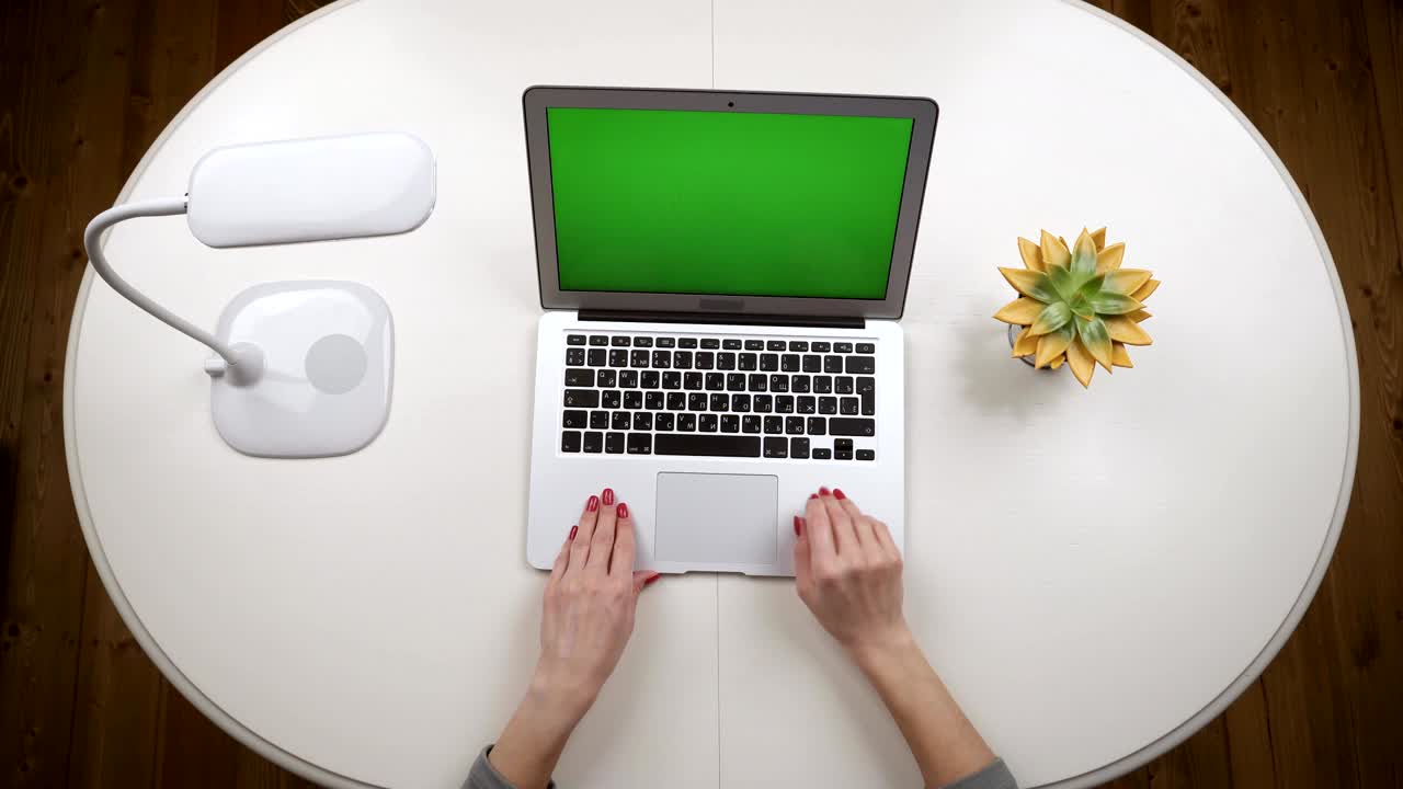 Ragazza freelance apre un laptop appoggiato su un tavolo con una lampada e dei fiori, digita del testo e lo chiude. Vista dall'alto. Mani in primo piano su chroma key.