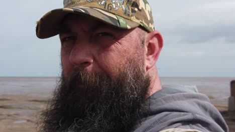 Lonely-bearded-urban-man-wearing-baseball-cap-looking-across-windy-beach-slow-motion