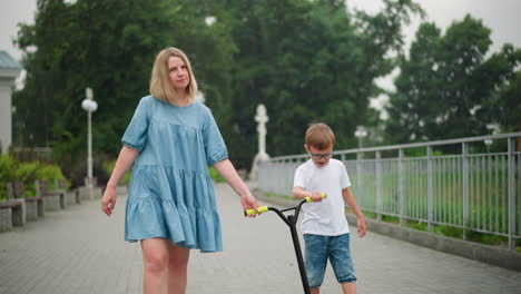a mother and her young son walk together on a peaceful path, the mother gently holds her son s hand while he pushes a scooter