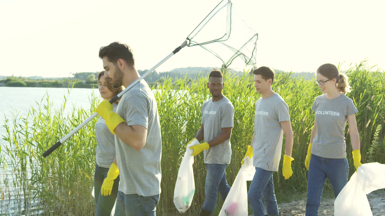 Environmental Young Mixed Races Volunteers Walking At The Lake Bank With Plastic Bags Full Of Trash And Talking After Working As Cleaners