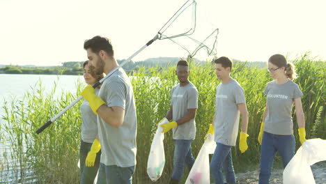 jóvenes voluntarios ambientales de razas mixtas caminando en la orilla del lago con bolsas de plástico llenas de basura y hablando después de trabajar como limpiadores