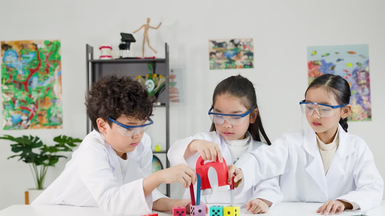 Tiga siswa sekolah dasar mengenakan kacamata pengaman dan jas lab sambil menjelajahi magnetisme dengan magnet tapal kuda