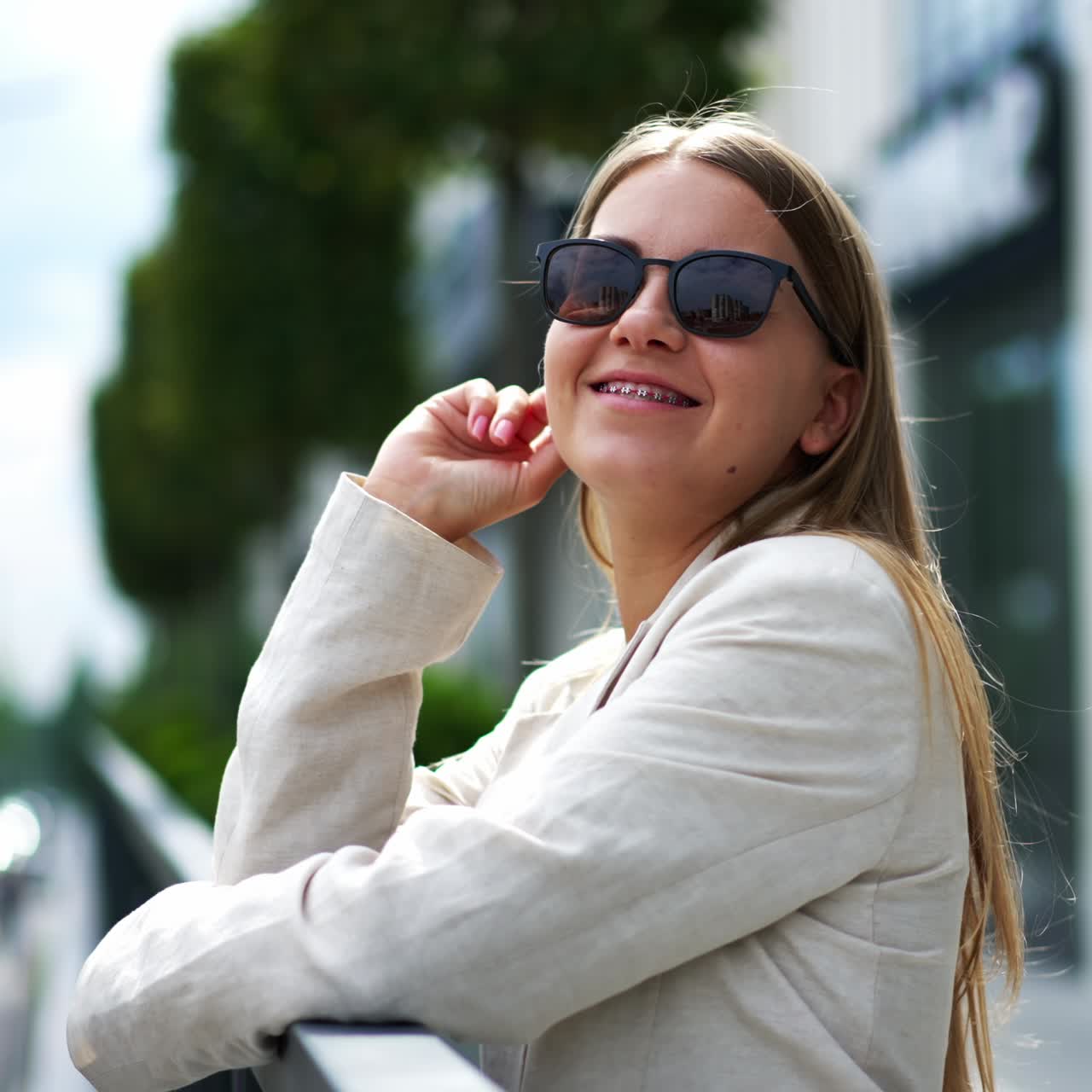 Happy young woman in sunglasses and white jacket in the street. Blonde takes off her glasses and smiles to the camera. Blurred backdrop