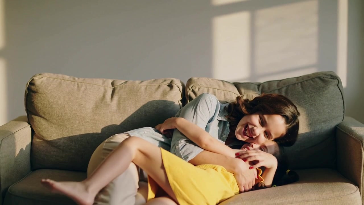 Running girl in yellow dress greeting mother in light blue shirt, sitting on sofa and cuddling