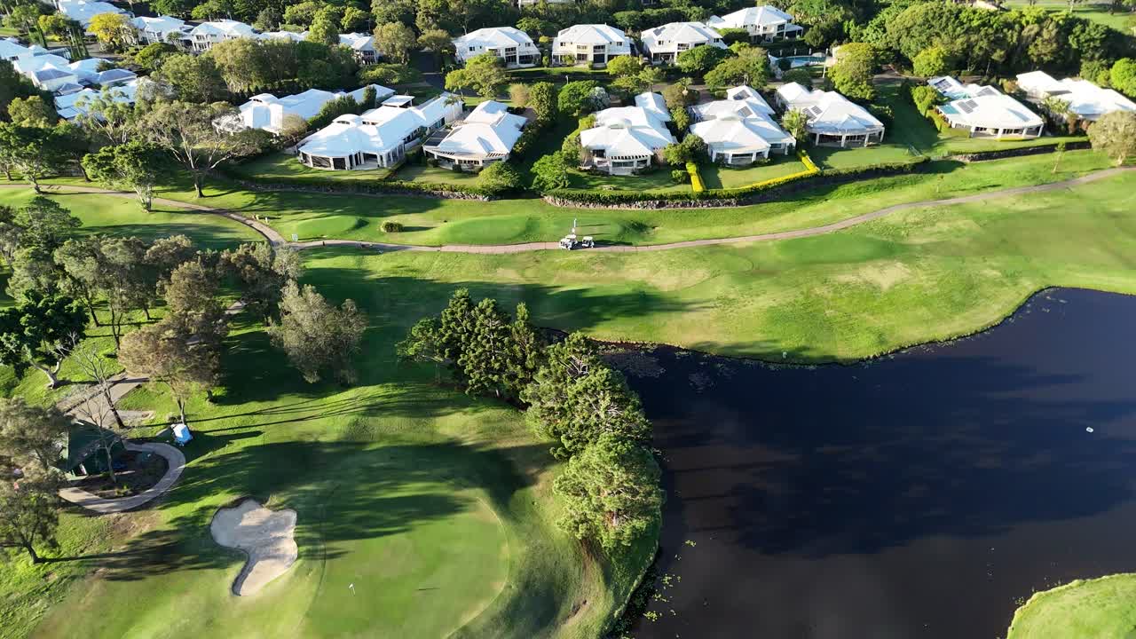 Drone footage captures a serene golf course with lush greenery, water hazards, and nearby residential areas during golden sunset hour