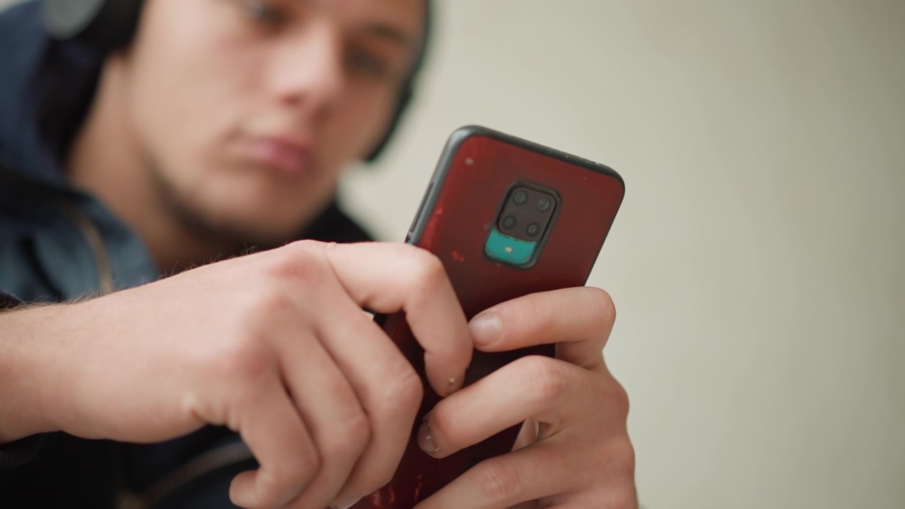 Close up of young man holding and interacting with red smartphone focusing on his hands tapping device. background is softly blurred emphasizing soft lighting and calm expression. Hands are