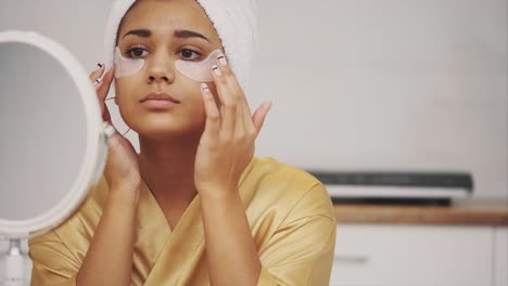 closeup portrait of beautiful woman after bath with towel on head puts patches under the eyes from wrinkles and dark circles. attractive female in bathrobe makes beauty treatments on the face at home.