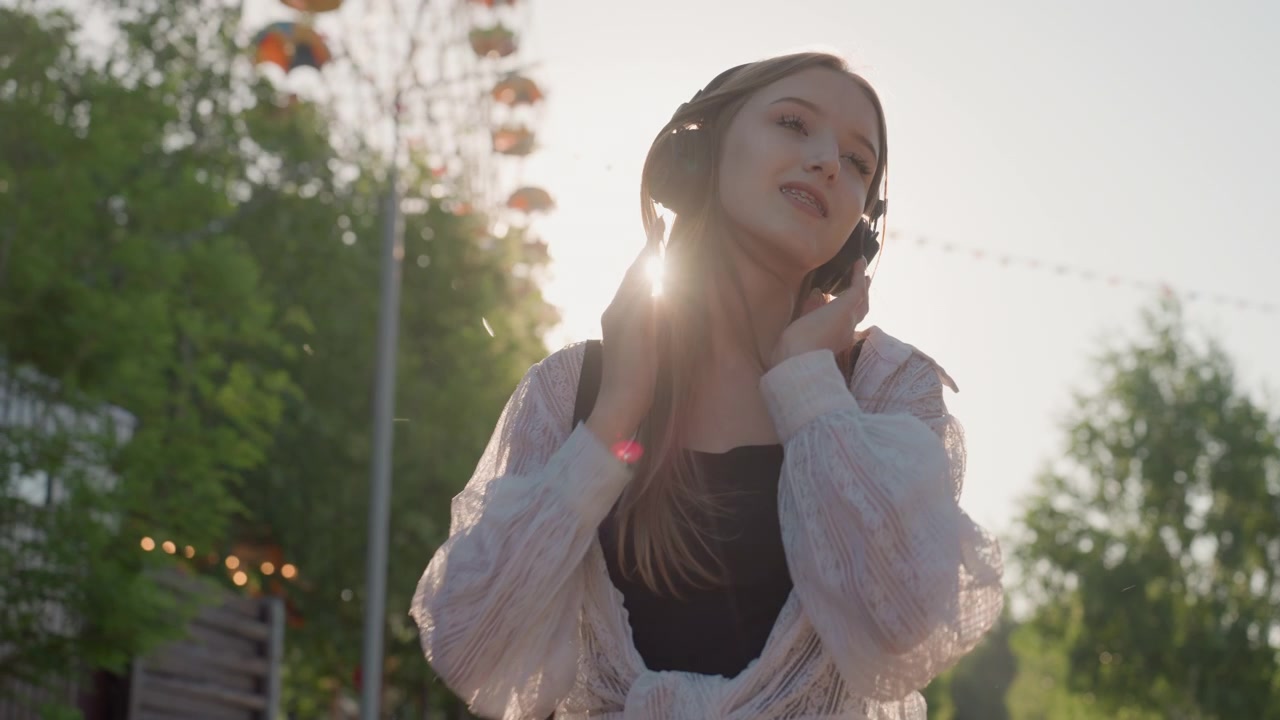 ヘッドホンを着用し、屋外で手のジェスチャーを交えながら音楽を楽しむ若い女性のクローズアップ。女性は長い髪で、明るい日差しの中、暗いトップスの上に明るい色のショールを羽織っている。