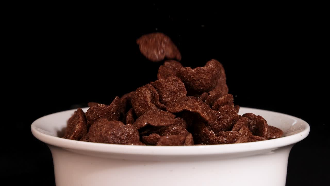 Chocolate cereal pieces drop into a white bowl against a black background, highlighting texture and motion