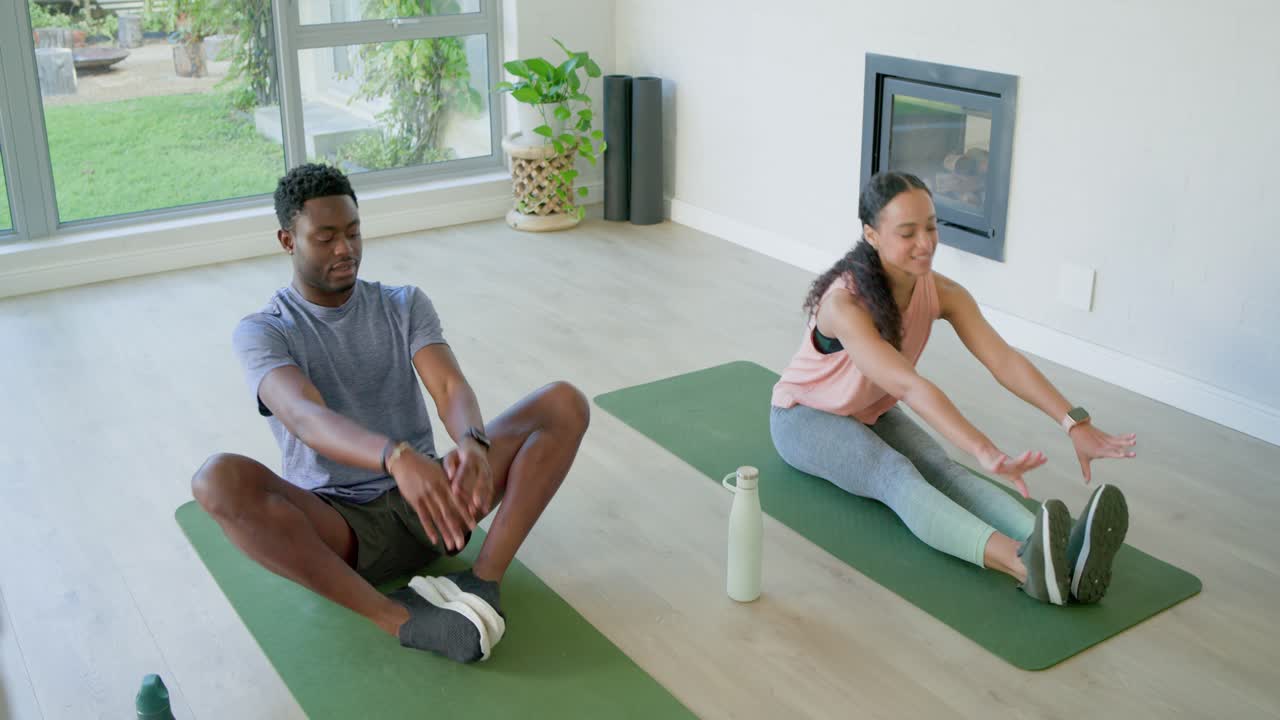 African American friends on green mats at home, woman leading stretching as man following, warming