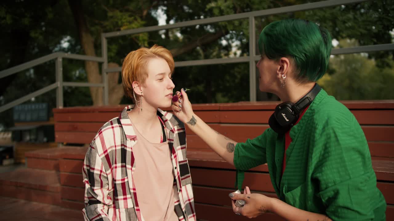 una chica con cabello verde en una camisa verde con auriculares hace maquillaje para su novia con cabello naranja brillante en una camiseta a cuadros en asientos marrones cerca del parque de skate en el verano en el parque