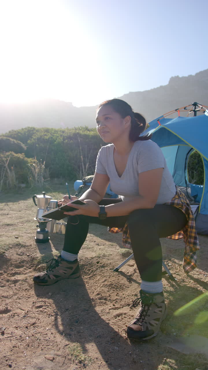Vertical video: Camping in mountains, woman sitting by tent holding coffee cup and relaxing