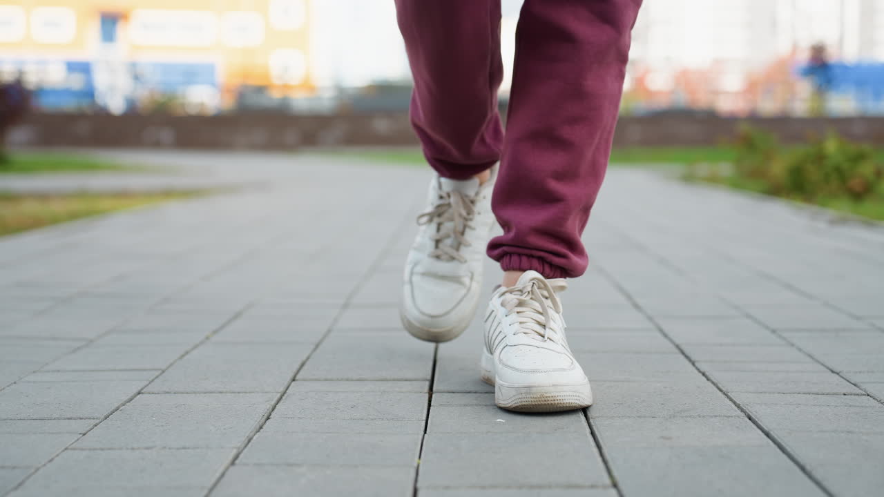 Close up leg view of athletic lady wearing white sneakers and burgundy jogger trousers walking on gray tiled pavement in urban park setting capturing stride motion and footwear textured detail