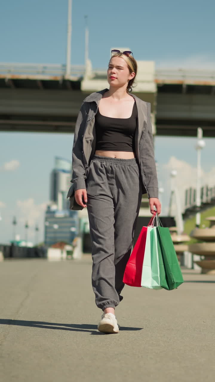 dama camina sosteniendo bolsas de compras en su mano derecha, con ropa gris abierta, la gente camina en el fondo cerca de un chaleco de flores, con un puente visible y coches conduciendo por encima