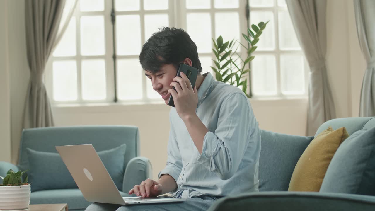 Asian Man Sitting Sofa In Living Room While Speaking By Phone And Working On Laptop, Video In 4K
