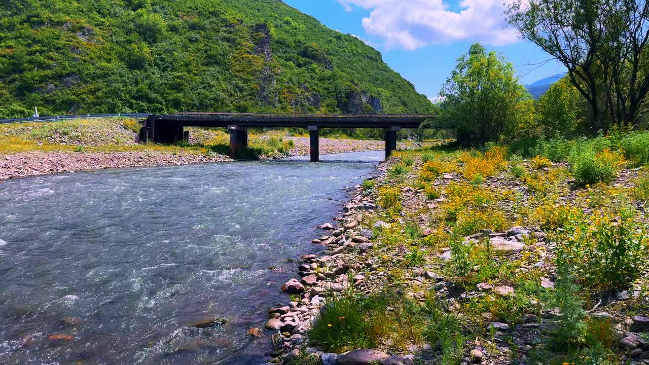 A Picturesque View of a Serene Riverbank Under a Clear Blue Sky, Framed by Lush Green Hills and a Sturdy Bridge Connecting Both Sides