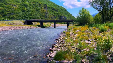 a picturesque view of a serene riverbank under a clear blue sky, framed by lush green hills and a sturdy bridge connecting both sides