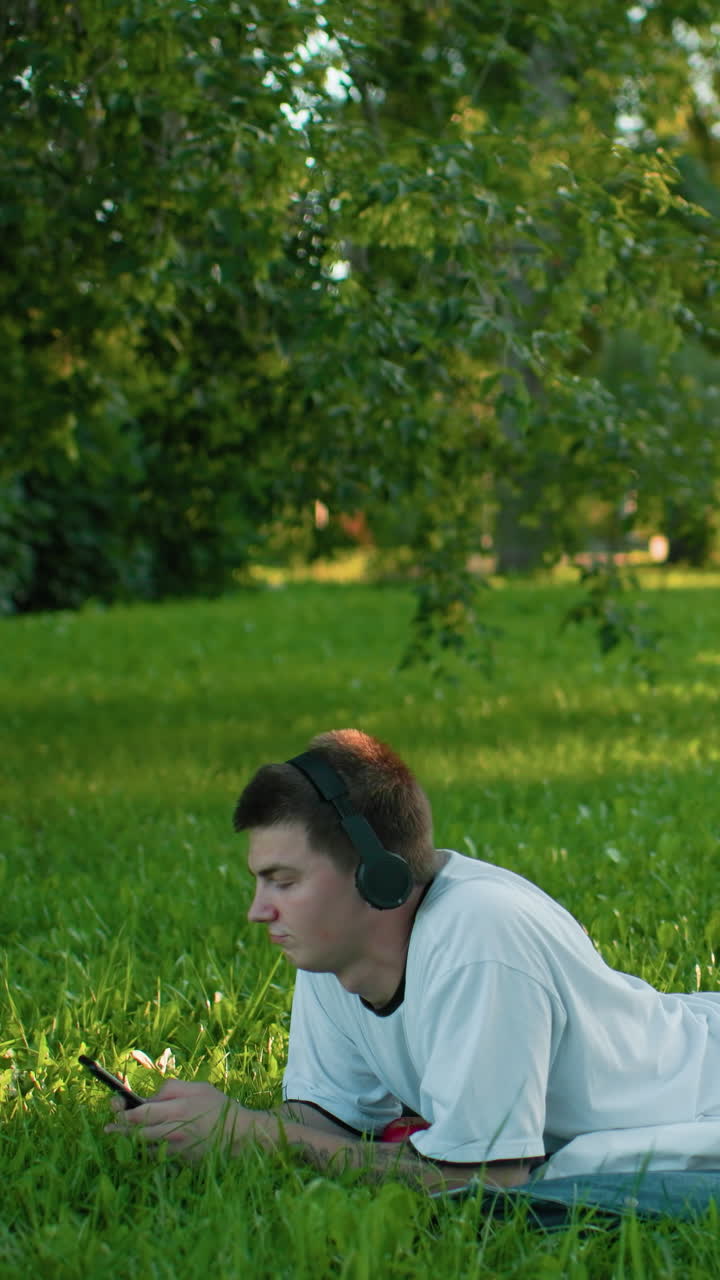 헤드폰을 달고 풀 위에 누워있는 젊은이, 휴대전화에 몰두하고, 울창한 초록색과 조용한 공원 환경에 둘러싸여, 그의 편안한 자세와 집중된 시선은 여가 시간을 반영합니다.