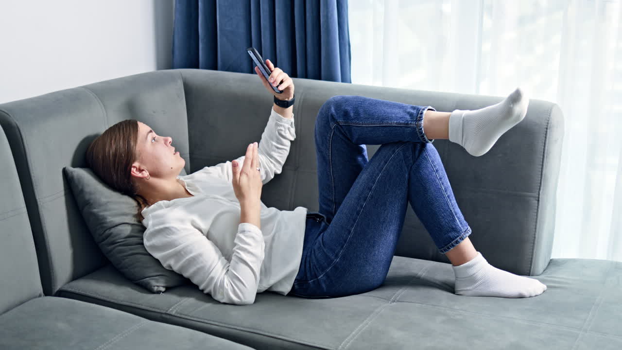 Donna felice seduta sul divano di casa, che scorre i social media sul telefono cellulare. Giovane donna che si rilassa sul divano, sentendosi intrattenuta, allegra, felice, godendosi il momento.