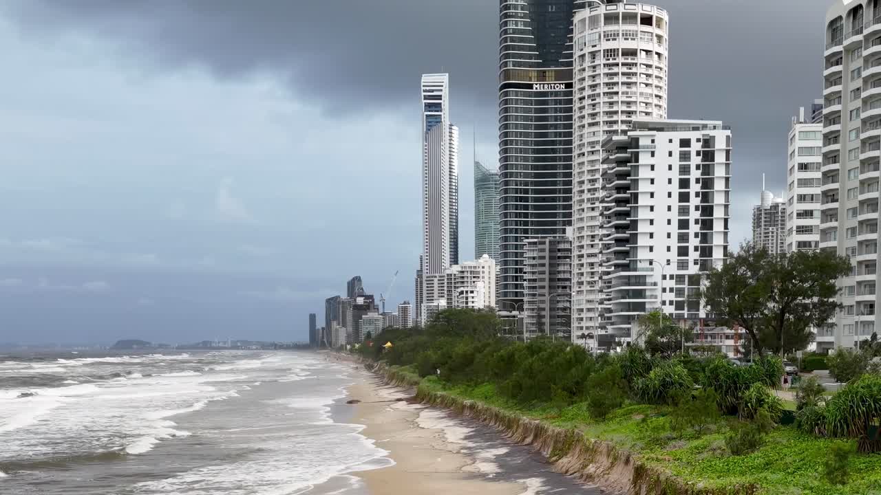 Aerial view of high-rise buildings along a stormy coastline with turbulent waves and overcast skies.