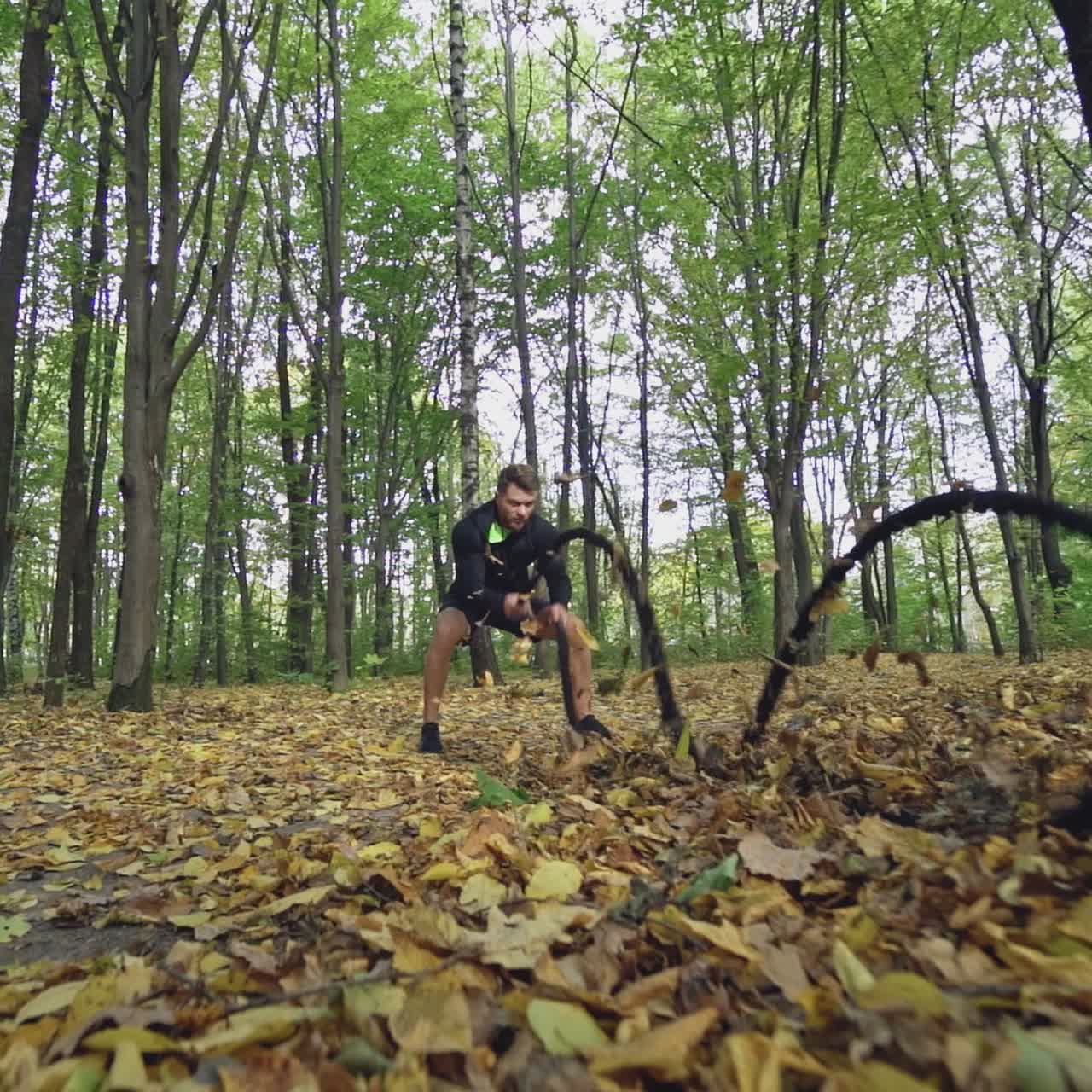 Sportsman doing waves with battle ropes in the autumn forest. Athletic man in black sportswear is training with ropes and goes away. Slow motion.