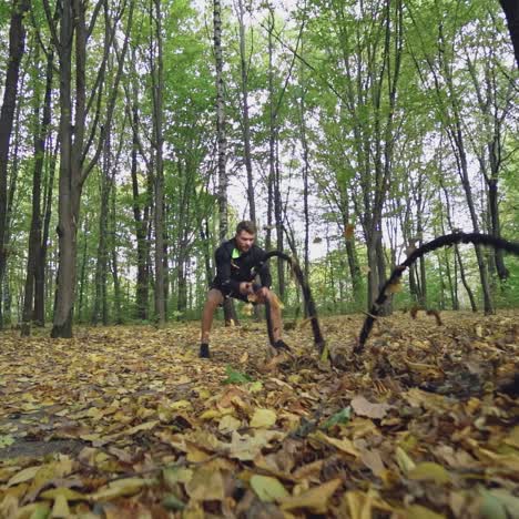 sportsman doing waves with battle ropes in the autumn forest. athletic man in black sportswear is training with ropes and goes away. slow motion.
