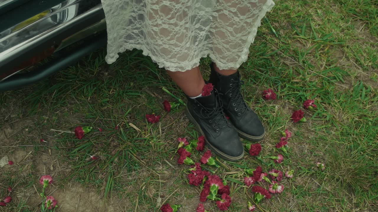 Woman in a lace dress sitting near a car with flowers around her boots