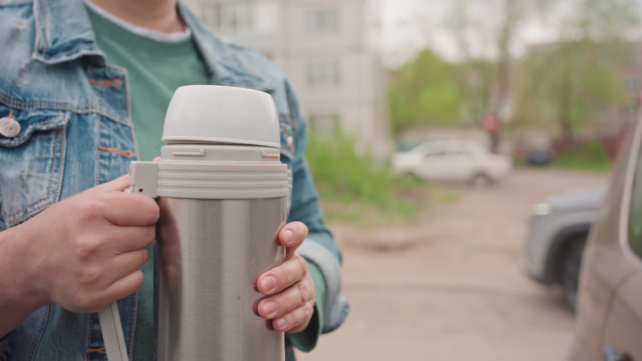 Caucasian Woman Opening Thermos Outdoors Denim Jacket Green Shirt Holding Metal Container Unscrewing Lid Scanning Residential Street Parked Cars Calm Morning Routine Offering Warm Beverage Neighborly