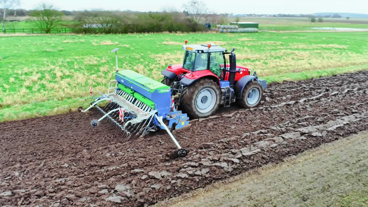 A 4k aerial view of a tractor pulling a seed drill on a wet field and sowing the next crop of wheat.
