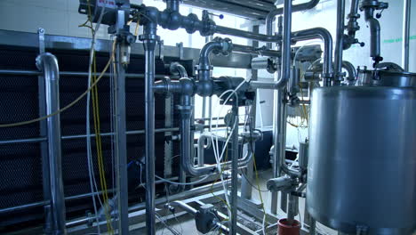 Stainless pipes and tubes as part of the equipment at modern dairy production. Metal tubes and big tank at the backdrop of large window. Silver pipes joined and crossed with each other.