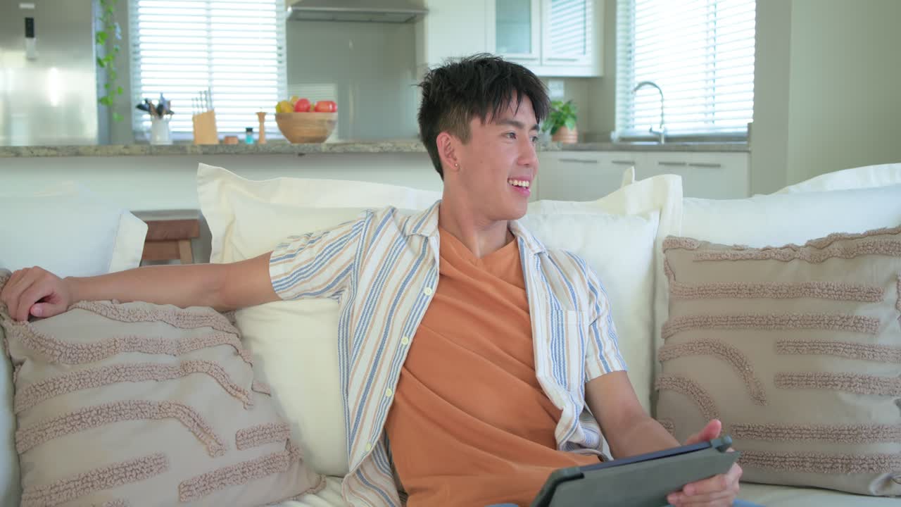 オレンジ色のTシャツを着たアジア人成人男性が自宅のソファに座り、タブレットをタップしながらコンテンツに笑顔を見せている。