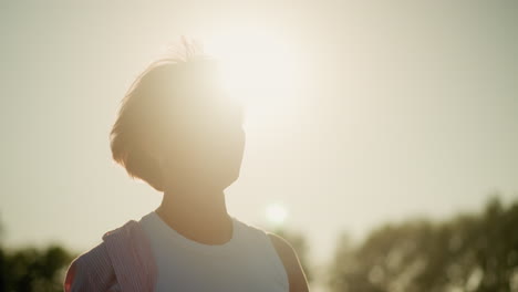 woman standing outdoors with bright sunlight radiating behind her, smiling warmly while looking at something in distance, sunlight creates halo effect around her head