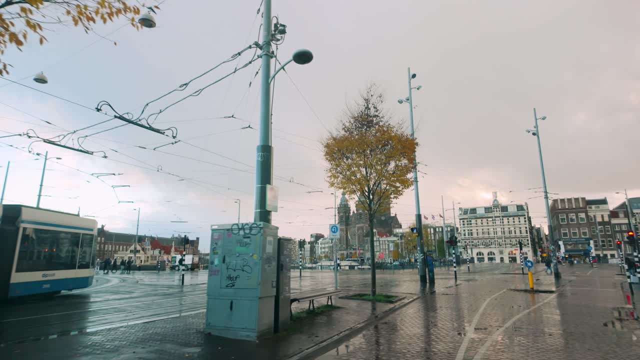 Amsterdam's Museumplein featuring a colorful advertisement pillar, tram tracks, wet pavement, autumn trees, and historic architecture under an overcast sky. Location: Amsterdam, Netherlands