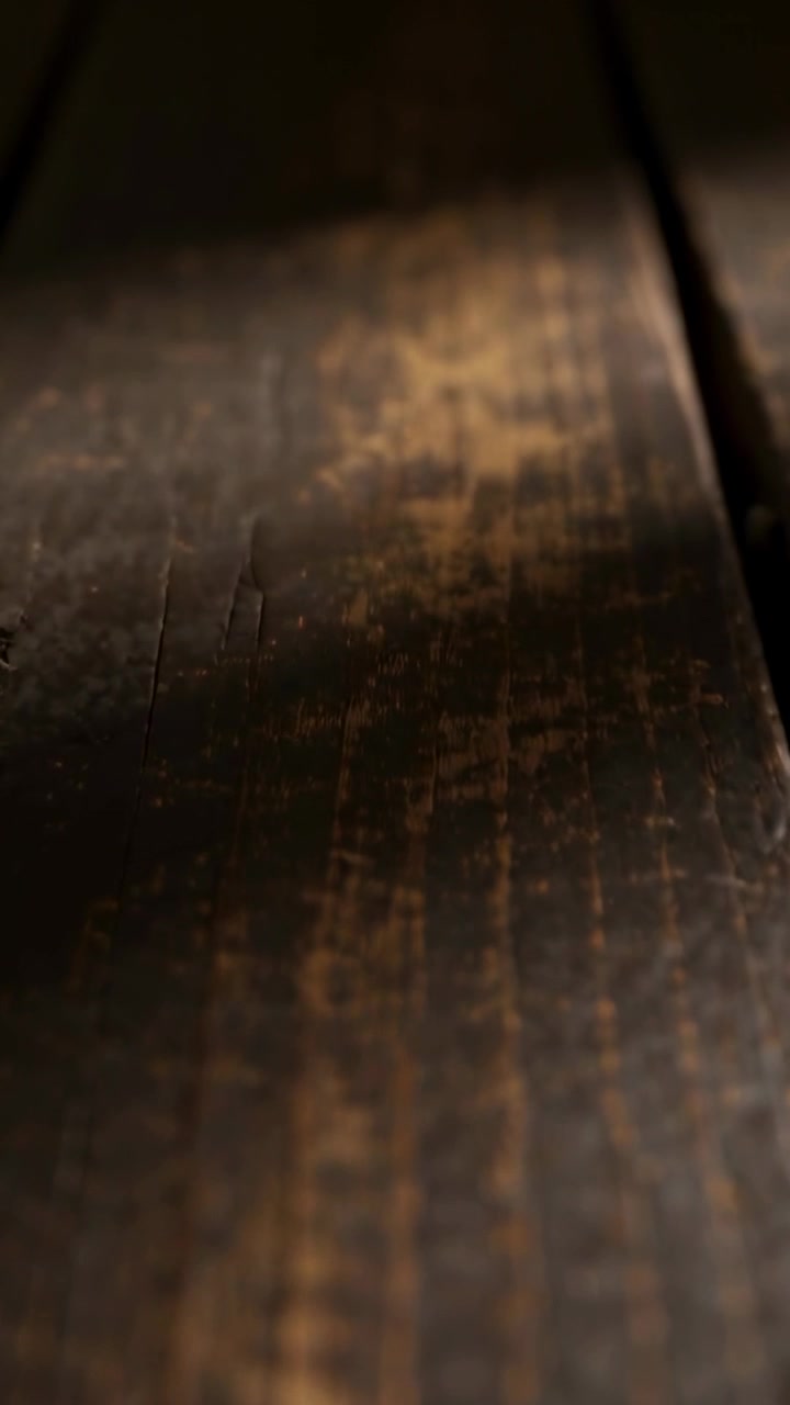 Video verticale: La telecamera si sposta con un'angolazione bassa, rivelando una tavola di legno usurata in una stanza in penombra, che mostra le venature.