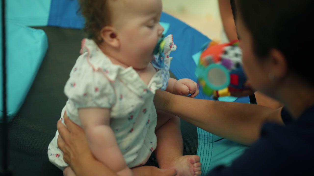 Bebê vestindo um macacão estampado e chupeta, deitado em um tapete azul, com um dos pais segurando gentilmente