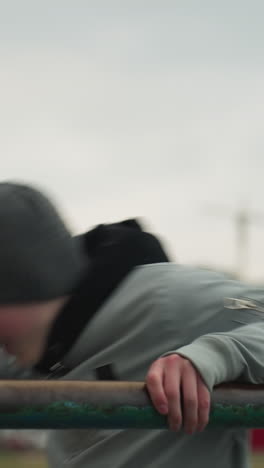 close-up of a young boy working out on an iron bar, wearing a gray sweater and beanie, the background features a blurred view of people and buildings