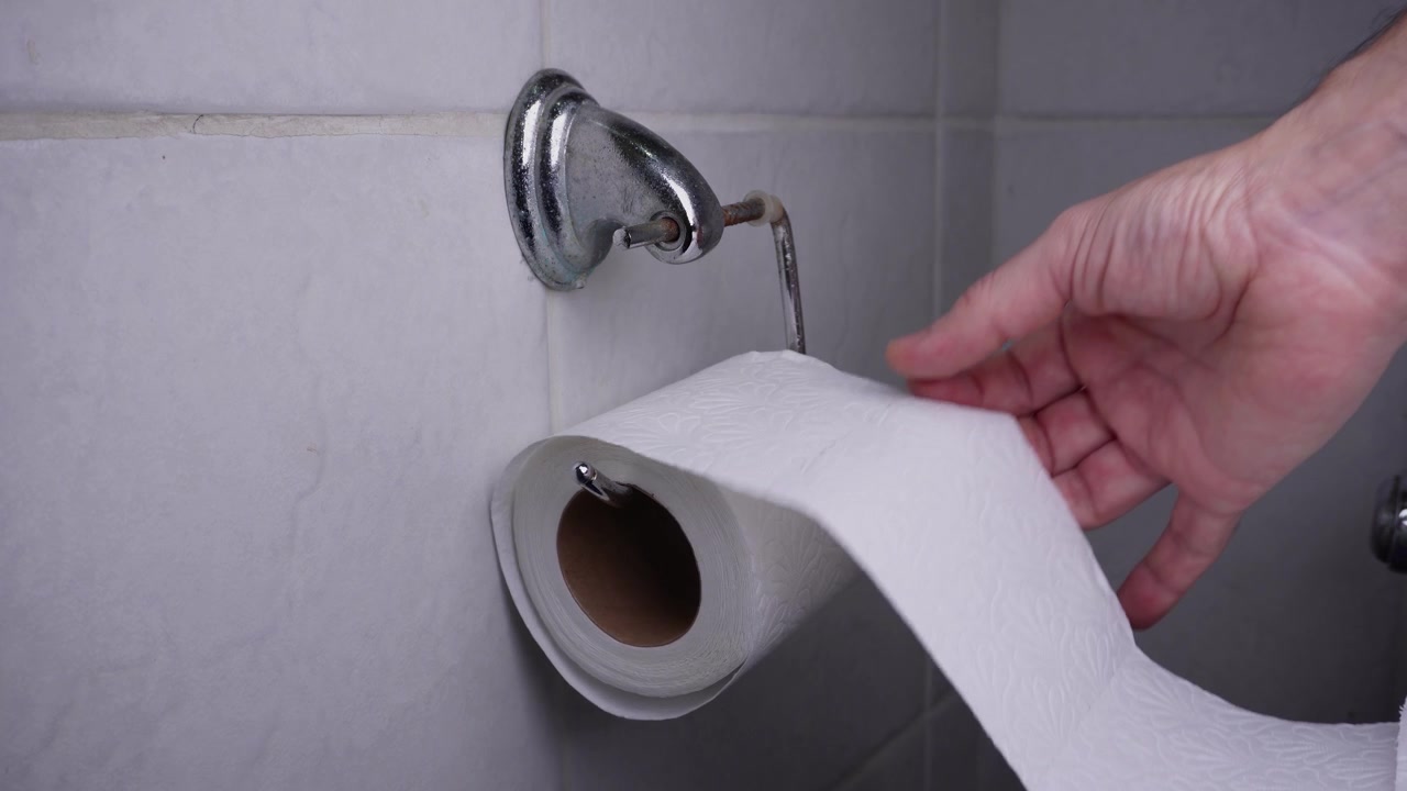 A hand comes into frame and starts pulling and unrolling a toilet paper roll thats on a metal holder on a bathroom wall.