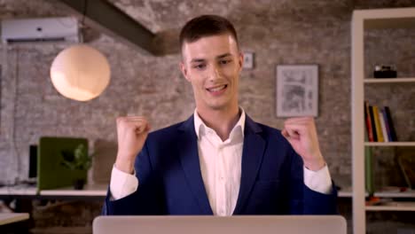 young concentrated businessman is watching laptop in office, happy because of good results, keeping hand fist, yes gesture, business concept, communication concept